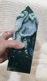 A green moss agate crystal point being held by a hand, with a blurred background suggesting a close-up view.