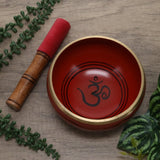 orangesinging bowl with Om symbol and wooden mallet on a wooden surface with green leaves.