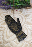 Decorative black palm with gold designs and an incense stick on a textured surface with greenery.