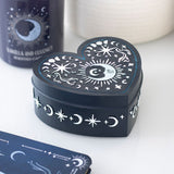 Heart-shaped black box with white celestial designs on a white surface with a candle and card in the background.