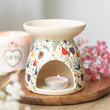 Decorative ceramic oil burner with a lit candle on a wooden surface, surrounded by flowers and a mug.