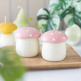 Two mushroom-shaped ceramic containers with pink caps on a wooden board with blurred greenery in the background.