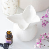 White ceramic diffuser with a bottle of essential oil and pink flowers on a light surface
