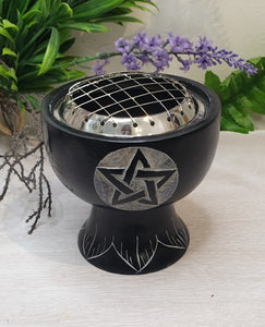 Black metal fire bowl with pentagram design on a white surface with plants in the background
