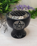 Black metal fire bowl with pentagram design on a white surface with plants in the background