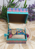 Colorful beaded bracelet with a heart charm in a decorative box on a wooden surface with flowers in the background.
