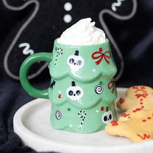Green mug with Halloween-themed design filled with hot chocolate and whipped cream, next to cookies on a dark background.