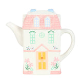 Ceramic teapot shaped like a house with a pink roof and yellow door on a white background