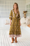 Woman wearing a patterned dress standing in a bright room with white walls and wooden floor.
