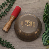 Tibetan singing bowl with a wooden mallet on a wooden surface with green leaves.
