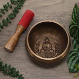 Bronze singing bowl with Buddha emblem and wooden mallet on a wooden surface with green plants.