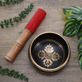 Black singing bowl with gold design and wooden mallet on a wooden surface with green leaves.