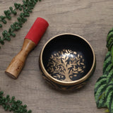 Decorative singing bowl with tree design on a wooden surface with a mallet and greenery.