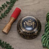 Decorative black and gold bowl with a wooden stick on a wooden surface with green leaves.