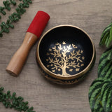 Black singing bowl with gold tree design and red mallet on a wooden surface with green leaves.