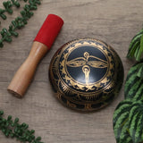 Decorative black and gold bowl with a wooden handle and red cushion on a wooden surface with green leaves.