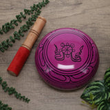 Pink singing bowl with a wooden handle on a wooden surface with green leaves.
