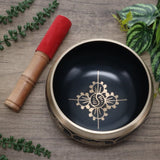 Decorative singing bowl with a red mallet on a wooden surface with green leaves.