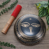 Decorative singing bowl with a wooden striker on a wooden surface with green leaves.