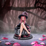 Figurine of a witch with pink hair sitting on a cauldron in a mystical setting with tree branches and petals.