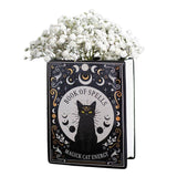 Decorative book with a black cat illustration titled 'Book of Spells: Magick Cat Energy' filled with white flowers on a white background.