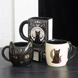 Three black cat-themed mugs on a dark surface with a book in the background.