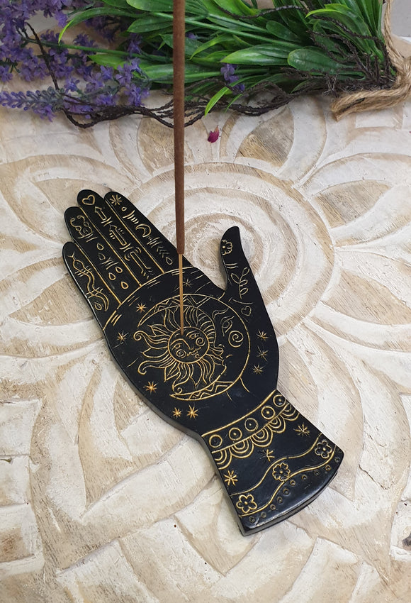 Decorative black palm with gold designs and an incense stick on a textured surface with greenery.