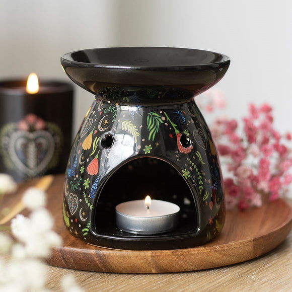 Decorative oil burner with floral designs on a wooden tray, surrounded by candles and flowers.