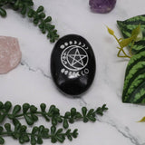 Black stone with a white pentagram symbol on a marble surface with greenery and crystals.
