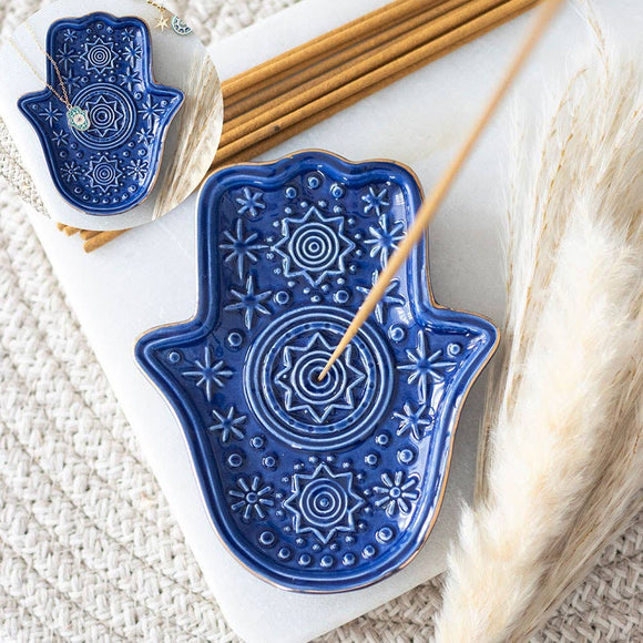 Blue Hamsa-shaped ceramic dish with decorative patterns on a white surface with incense sticks and dried plants.