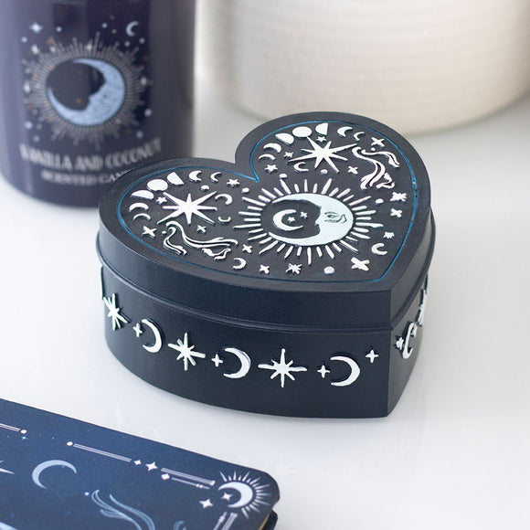 Heart-shaped black box with white celestial designs on a white surface with a candle and card in the background.