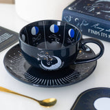 Black ceramic cup and saucer with moon phases design on a white surface.
