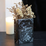 Decorative book-shaped vase with 'Book of Spells' design filled with dried flowers on a dark surface.