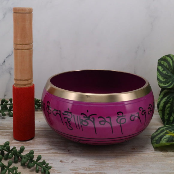 Pink singing bowl with gold rim and wooden striker on a wooden surface with green leaves.