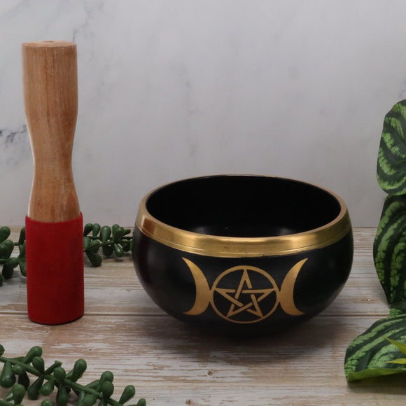 Black singing bowl with gold pentacle design and wooden mallet on a wooden surface with greenery.