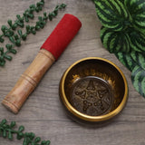 Brass singing bowl with pentagram design and wooden mallet on a wooden surface with green leaves.