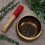 Bronze singing bowl with decorative design on a wooden surface with green leaves.