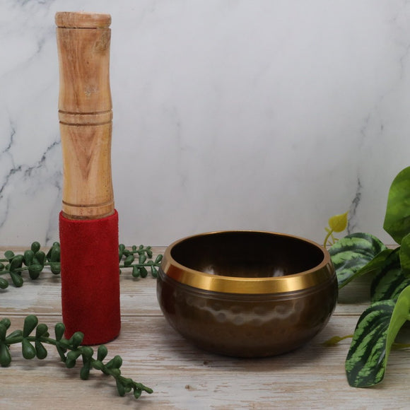 Brown singing bowl with gold rim and wooden mallet on a marble surface with greenery