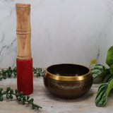 Brown singing bowl with gold rim and wooden mallet on a marble surface with greenery