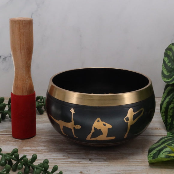 Black singing bowl with gold yoga pose designs on a wooden surface with a wooden mallet.