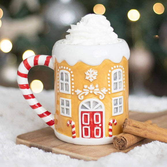 Gingerbread house mug with candy cane handle on a snowy surface with blurred Christmas lights in the background.