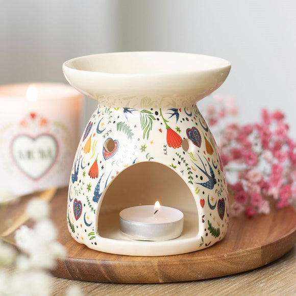 Decorative ceramic oil burner with a lit candle on a wooden surface, surrounded by flowers and a mug.