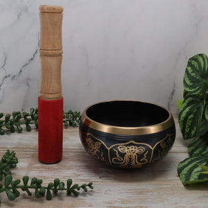 Black singing bowl with gold patterns and a wooden mallet on a marble surface with green plants.