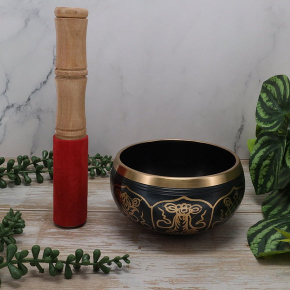 Black singing bowl with gold patterns and a wooden mallet on a marble surface with green plants.