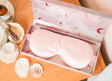 Pink eye mask in a box on a wooden surface with seashells and a bottle.