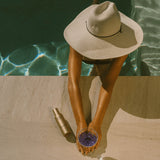 Person in a sun hat by a pool holding a glass with a bottle next to them.