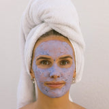 Woman with a blue facial mask on her face and a white towel wrapped around her head.