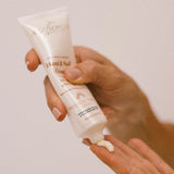 Hand holding a tube of Mira Loma Hand & Nail Cream with visible cream on the fingers against a neutral background.