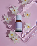 A bottle of essential oil with a purple label surrounded by white flowers on a pink background