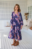 Woman wearing a floral dress standing on a wooden floor with a white wall and decorative items in the background.